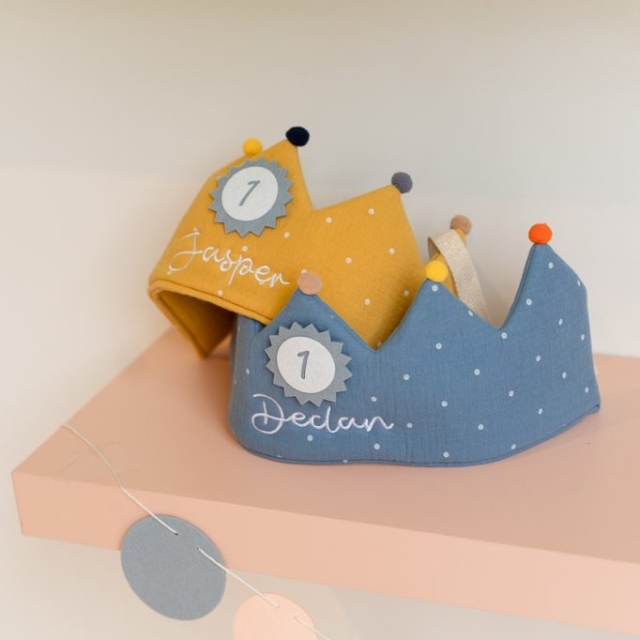 Two fabric birthday crowns, one yellow labeled "Jasper" and one blue labeled "Declan," each with a "1" badge, rest on a pink shelf above a garland of paper circles.