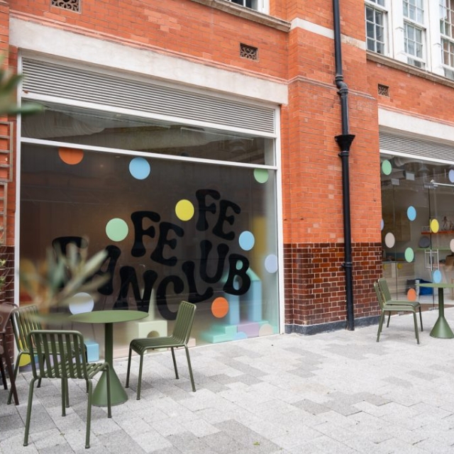 Outdoor tables and chairs are set up on a sidewalk in front of a brick building with large windows featuring colorful polka dots and bold "FE FAN CLUB" lettering.
