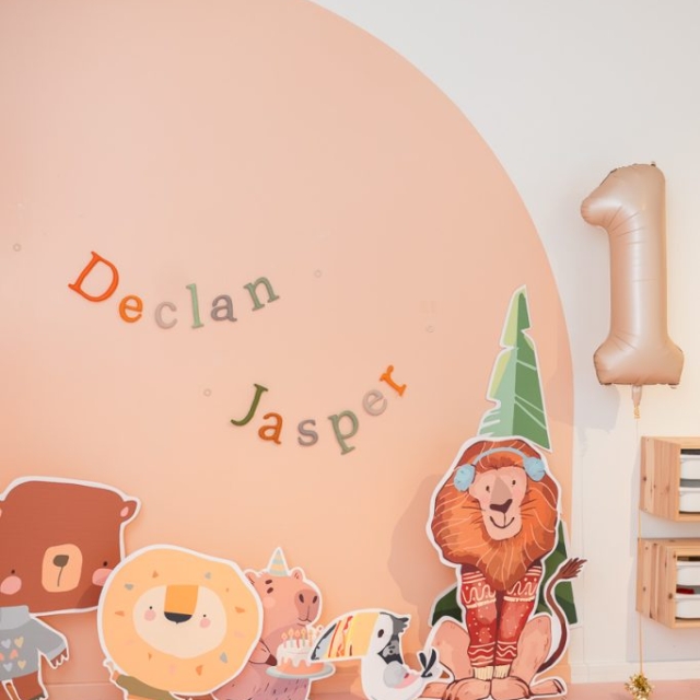 A pastel-colored birthday setup features a "Declan Jasper" banner, animal cutouts wearing party hats, and a large gold balloon in the shape of the number one.