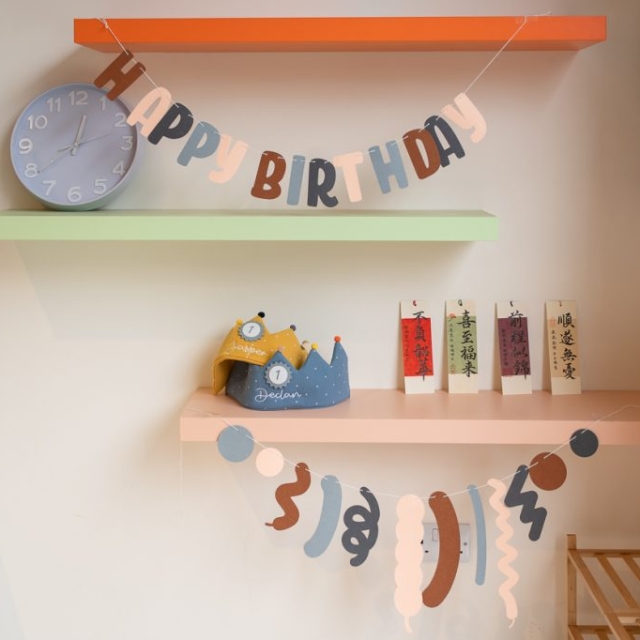 A wall with three shelves displays a clock, birthday decorations, a small yellow crown, and several vertical cards with Chinese characters.