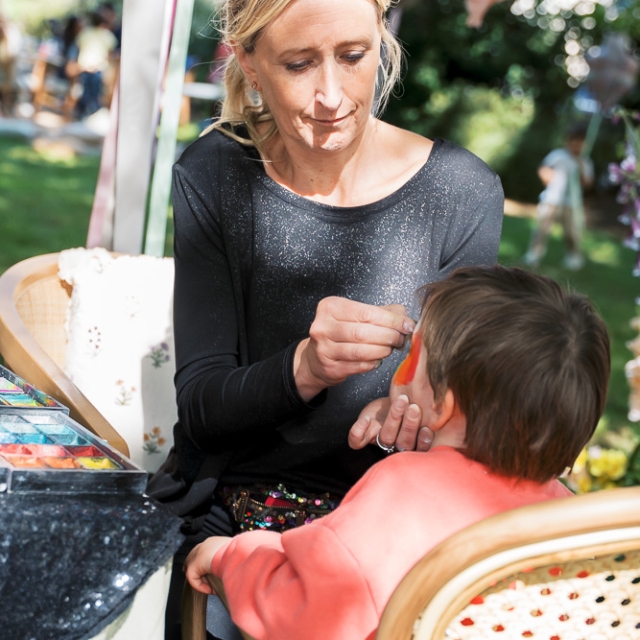 5th birthday garden party – Elisa Design Photography (1) A woman applies face paint to a young child sitting in a chair outdoors, with art supplies on the table and greenery in the background.