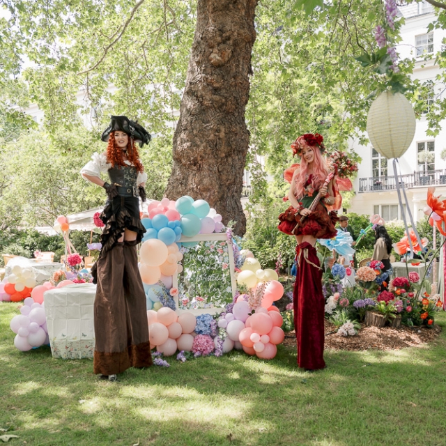 5th birthday garden party – Elisa Design Photography (2) Two people on stilts in elaborate costumes stand on grass near a large tree, with colorful balloons and decorations in a garden setting.