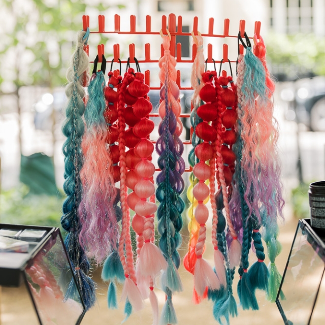 5th birthday garden party – Elisa Design Photography (3) A display rack holds colorful synthetic hair braids and ponytails in various bright shades, arranged outdoors on a sunny day.