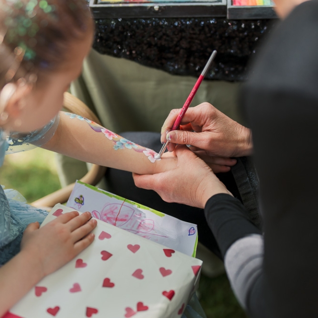 5th birthday garden party – Elisa Design Photography (4) An adult paints colorful designs on a child’s arm while the child holds a heart-patterned gift. A palette of paints is visible on the table.