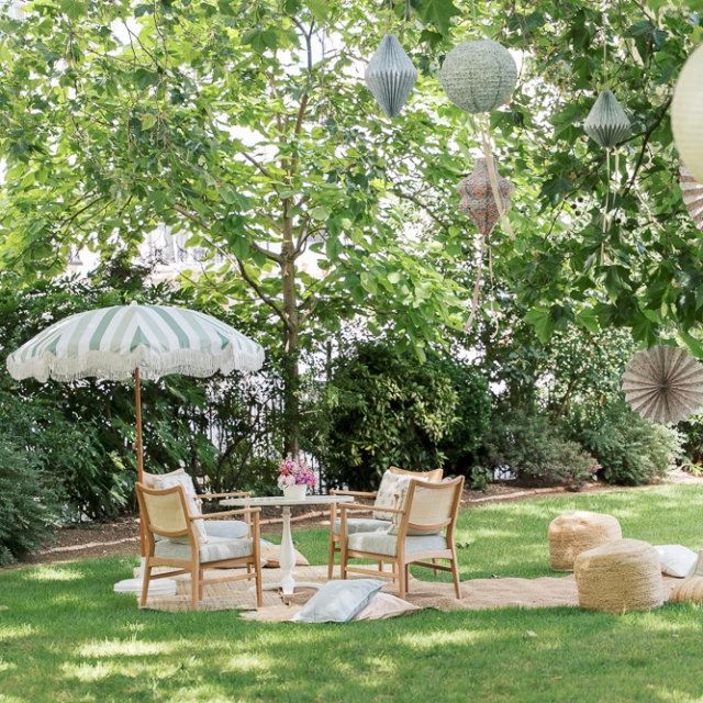Belgravia-5th birthday party – Elisa Design Photography Outdoor seating area with four chairs, a striped umbrella, a small table, poufs, and hanging paper lanterns on a grassy lawn under leafy trees.