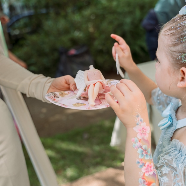 Belgravia-birthday party -photographer (1) A child with floral face paint reaches for a plate of pink desserts handed by an adult at an outdoor event.