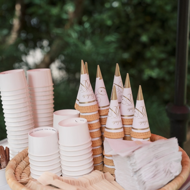 Belgravia-birthday party -photographer (10) A wicker basket holds stacked paper cups, cone-shaped wrappers on ice cream cones, wooden utensils, and napkins on a white surface, with greenery in the background.