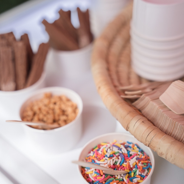 Belgravia-birthday party -photographer (11) White bowls with rainbow sprinkles, crushed nuts, and chocolate sticks are arranged on a table with stacked paper cups and wooden utensils.