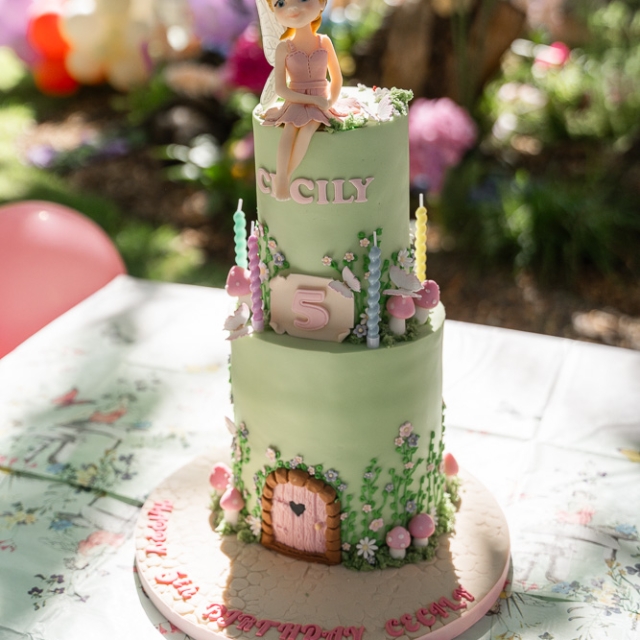 Belgravia-birthday party -photographer (13) Two-tier green birthday cake with pink flowers, a fondant door, the name “Cicily,” a number 5, and a doll topper, displayed on a decorated table outdoors.