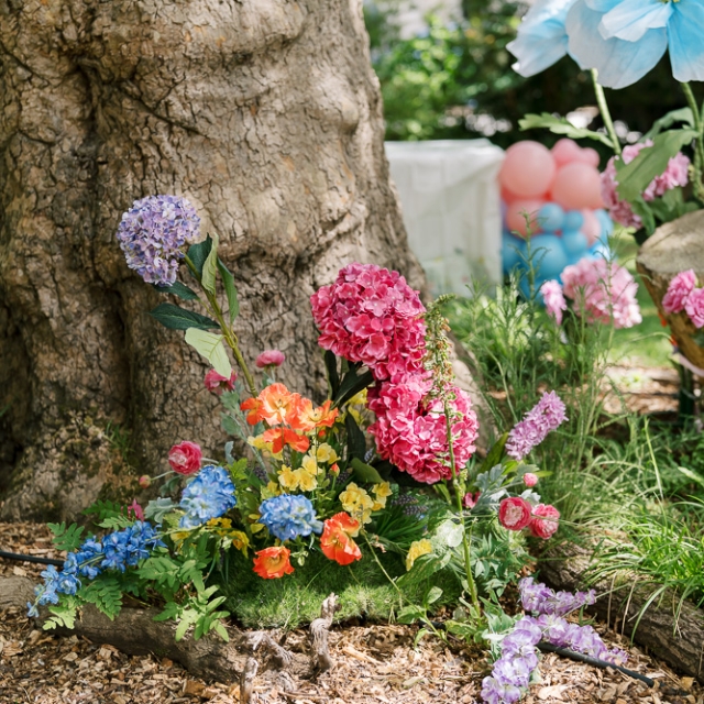 Belgravia-birthday party -photographer (3) Colorful artificial flowers are arranged at the base of a large tree in a garden, with balloons and greenery visible in the background.