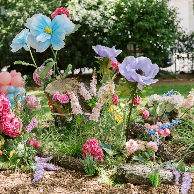 Belgravia-birthday party -photographer (4) A garden scene with oversized artificial flowers in bright colors, surrounded by greenery, tree trunks, and a cluster of pastel balloons in the background.