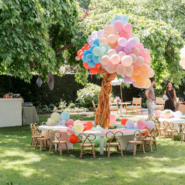 Belgravia-birthday party -photographer (5) Round tables and small chairs are arranged on grass with a centerpiece made of twisted balloons under large trees at an outdoor event.