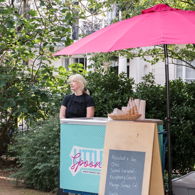 Belgravia-birthday party -photographer (9) A woman stands behind an ice cream cart with a pink umbrella. A chalkboard lists flavors including hazelnut, Oreo, salted caramel, raspberry coulis, mango sorbet, and vanilla.