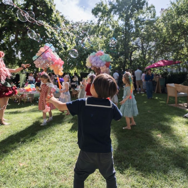 Belgravia-garden-party Children play with bubbles and balloons at an outdoor garden party, while adults look on under sunny, leafy trees.