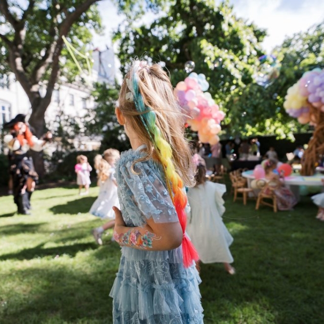 Belgravia-kids-party-photographer Children in colorful costumes play on a grassy lawn at an outdoor party with balloons, tables, and trees in the background.