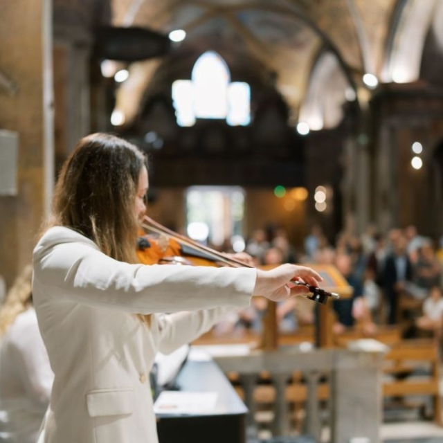 Baptisme-photographer-london-elisa-design-photography Person in a white jacket plays violin inside a church during a battesimo, with blurred audience seated in the background.