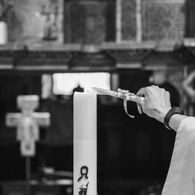 Christening-photographer-London2 A person lights a tall decorated candle with a taper in a church setting, symbolizing baptism; a cross and altar are visible in the background.
