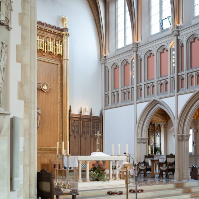london-Christening-photographer (16) Interior of an English church with tall arched windows, ornate wooden altar, marble flooring, candles, and chairs arranged for a baptism service.