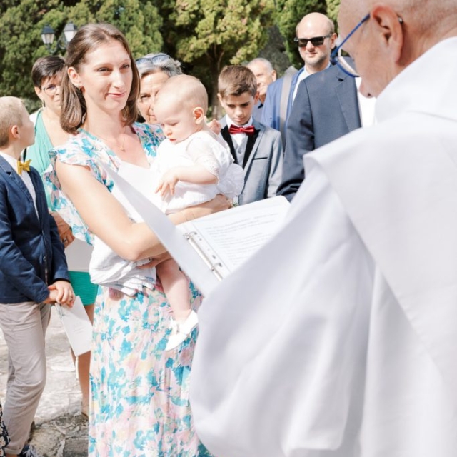 photographe-bapteme-Elisa-Design-Photography (13) A woman holding a baby stands among a group of people outdoors as a priest in white robes reads from a document, marking the special occasion of a baptism.
