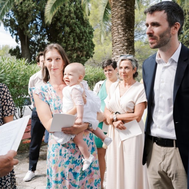 photographe-bapteme-Elisa-Design-Photography (18) A group of adults and a baby gather outdoors near palm trees, likely celebrating a baptism. A woman holds the baby, who appears upset, while an older man holds documents and addresses them during the battesimo ceremony.