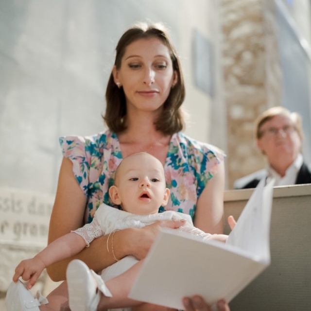 photographe-bapteme-Elisa-Design-Photography (19) A woman in a floral dress holds a baby dressed in white, with both looking at an open booklet in what appears to be a church or chapel setting, capturing a tender moment during a battesimo (baptism) ceremony.
