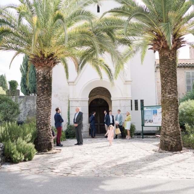 photographe-bapteme-Elisa-Design-Photography (3) A group of people stand and talk outside a white church with a bell tower, framed by two tall palm trees on a sunny day, perhaps gathered to celebrate a baptism.