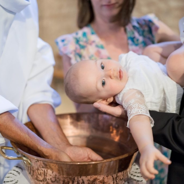 photographe-bapteme-Elisa-Design-Photography (31) An infant in a white outfit is held over a copper baptismal font by adults during a baptism ceremony, also known as battesimo in English.