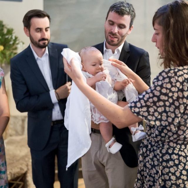 photographe-bapteme-Elisa-Design-Photography (36) Three adults stand around a baby dressed in white during a baptism ceremony, with one woman holding a cloth near the baby’s head.