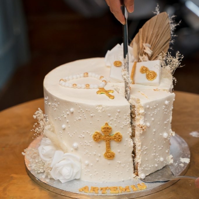 religious-event-photographer-england (20) A hand cuts a white decorated cake with gold crosses and white flowers on a round table, capturing the elegance of a baptism celebration. The word “ARTEMIUS” is written on the cake’s base, making this battesimo moment truly special.