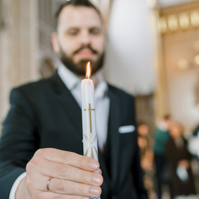 religious-event-photographer-england (45) A man in a suit holds a lit white candle with a gold cross and decoration inside a church, symbolizing the sacred ceremony of baptism.